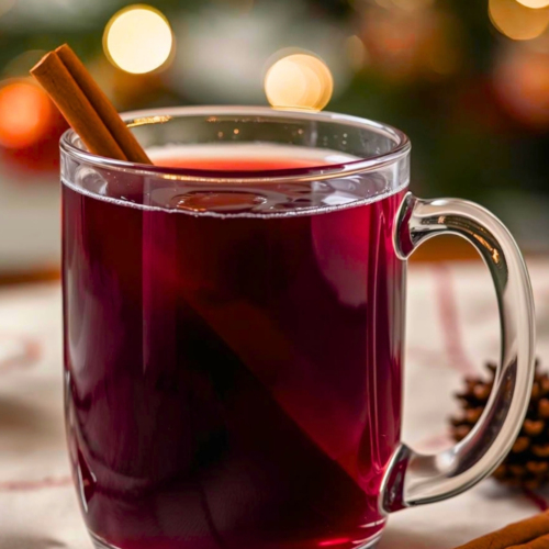 Cozy glass cup of red mulled wine with a cinnamon stick as garnish. Christmas tree in the background. Recipe for how to make the hot mulled wine.