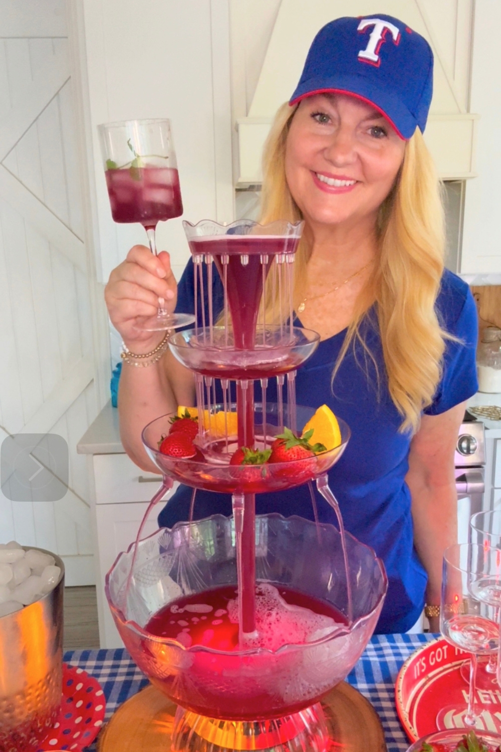 16 Big Batch Party Drink Recipes 2 Women wearing ball cap holding wine glass with a drink next to tall drink fountain with red punch flowing down.