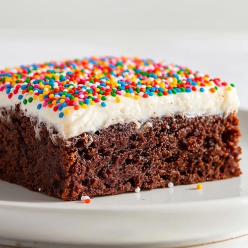 A plate with a Dot Cake Brownie. Recipe for brownies with thick coat of vanilla frosting and rainbow sprinkles.