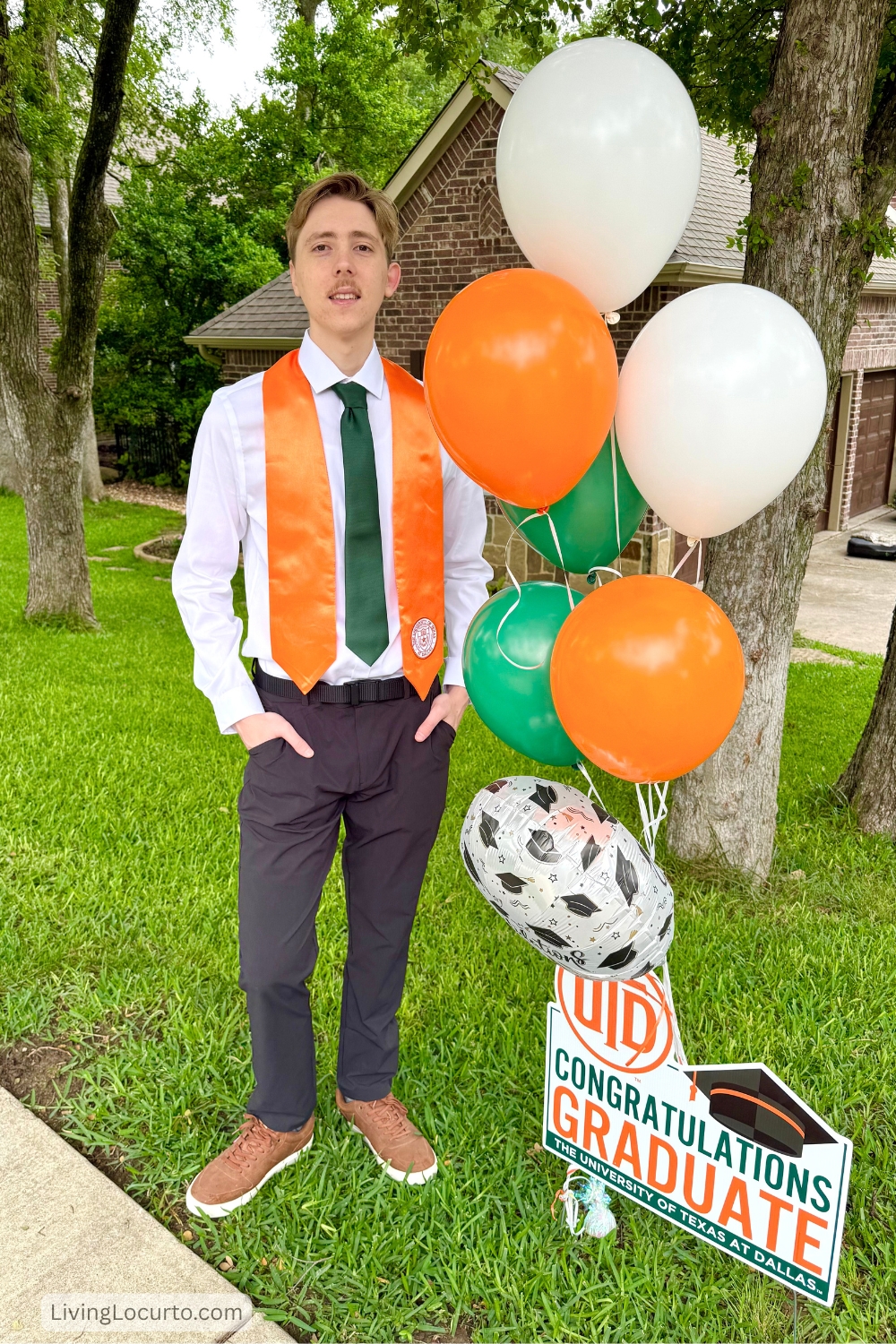 Young man in a tie standing next to a graduation sign with balloons in yard.